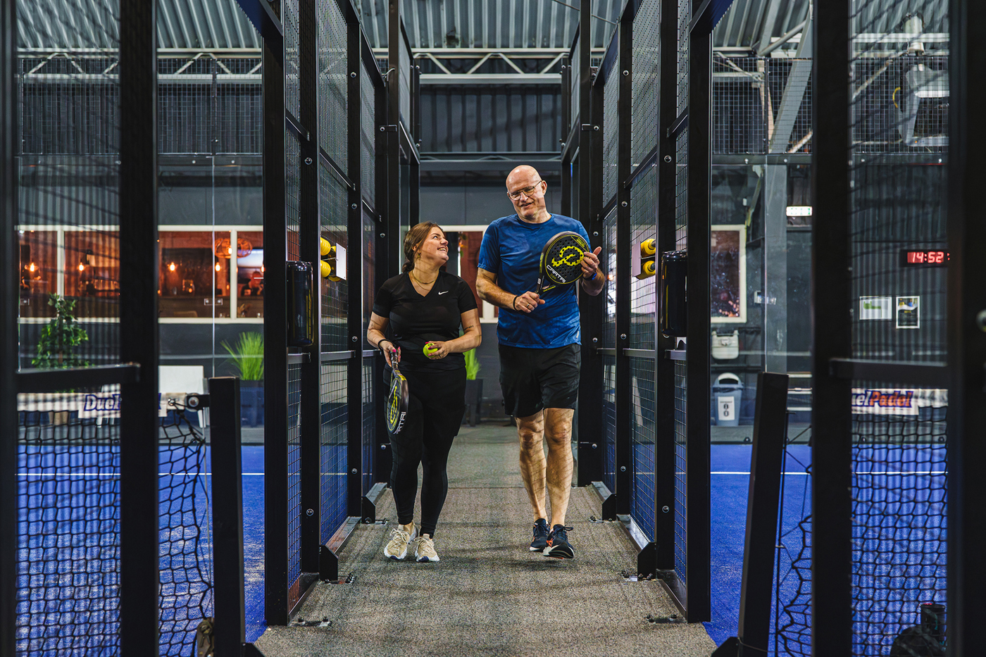 Man en vrouw lopen met padelrackets tussen indoor padelbanen