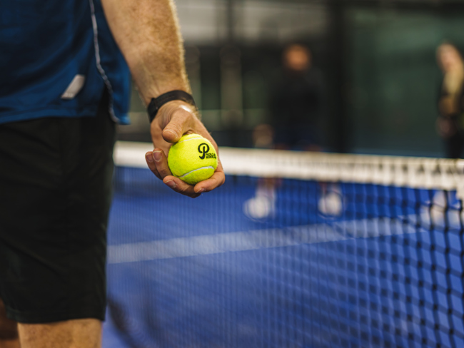 Close-up van een hand met een gele padelbal naast het net van een blauwe indoor padelbaan.