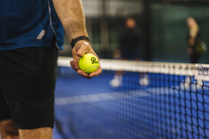 Close-up van een hand met een gele padelbal naast het net van een blauwe indoor padelbaan.