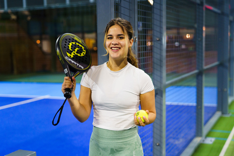 Een vrouw staat lachend naast een blauwe indoor padelbaan en kijkt recht richting de camera. Ze draagt een wit sportshirt en een lichtgroene sportbroek. In één hand houdt ze een zwart-geel padelracket omhoog en in haar andere hand een gele padelbal. 