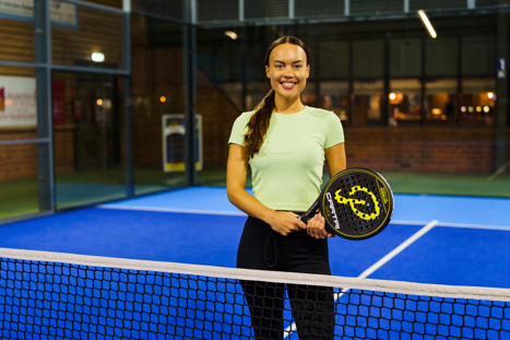 Vrouw in sportkleding staat lachend met een padelracket in haar hand op een blauwe indoor padelbaan.